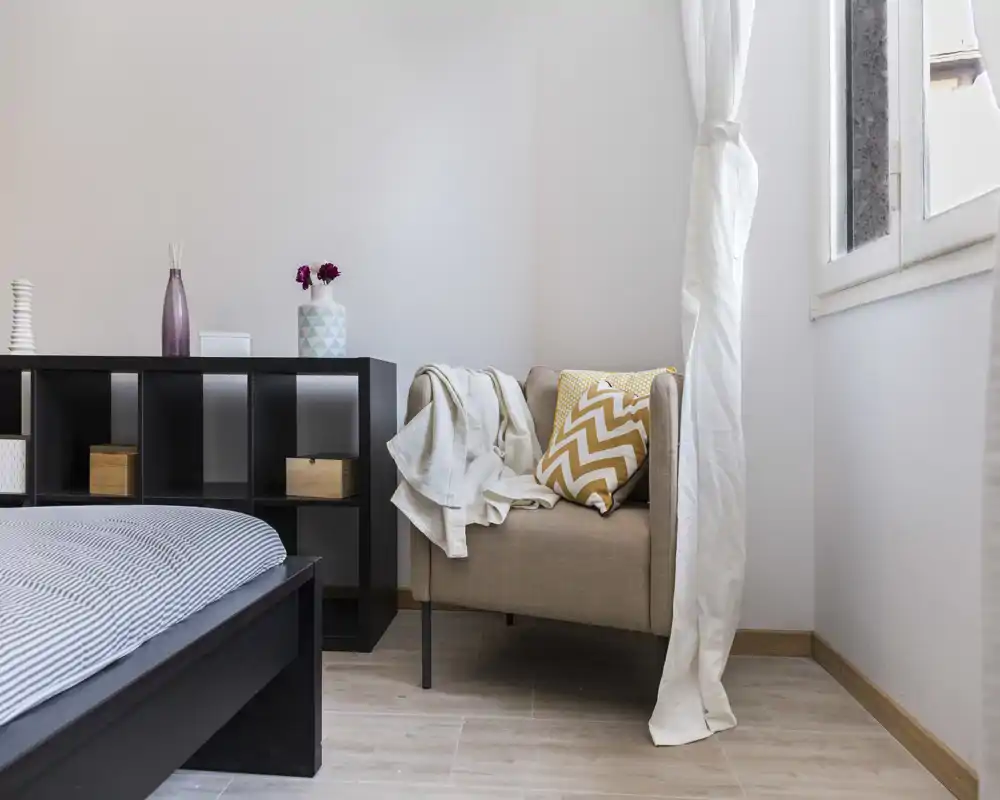 Angolo di camera luminoso e curato con telaio del letto visibile e una poltrona accogliente con cuscino a zigzag e plaid leggero.