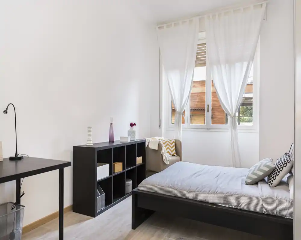 Camera da letto luminosa e minimale con letto ben rifatto vicino a una grande finestra, scaffalatura semplice e una piccola scrivania—stile moderno e molta luce naturale.