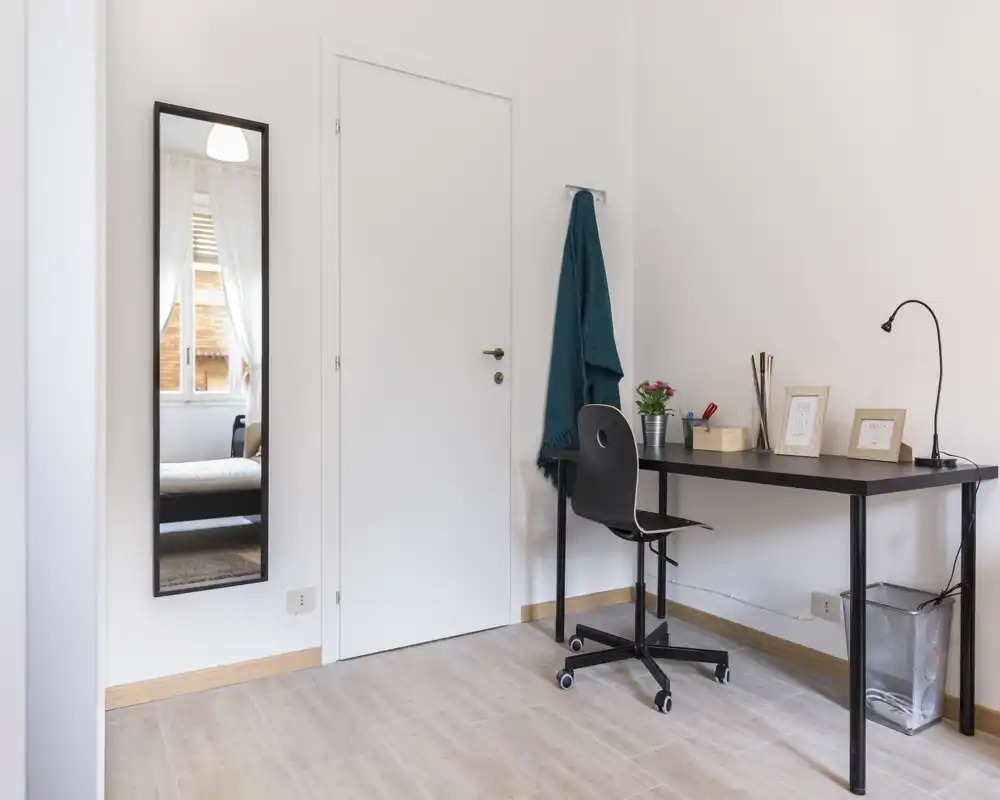 Angolo studio compatto e luminoso con una scrivania nera, sedia girevole, lampada da scrivania e cornici decorative; uno specchio a figura intera e un appendiabiti aggiungono funzionalità.