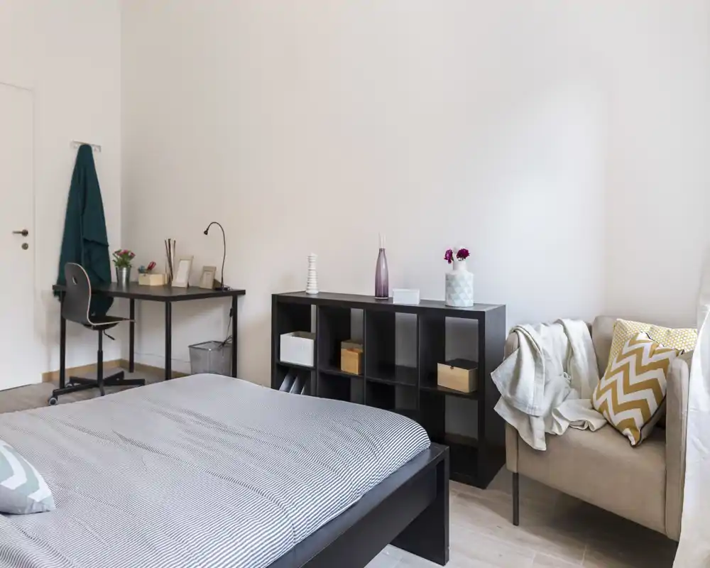 Ampia e luminosa camera da letto con un grande letto, cuscini decorativi e una poltrona accogliente. Un mobile a cubi nero e una piccola scrivania offrono spazio di archiviazione e lavoro.