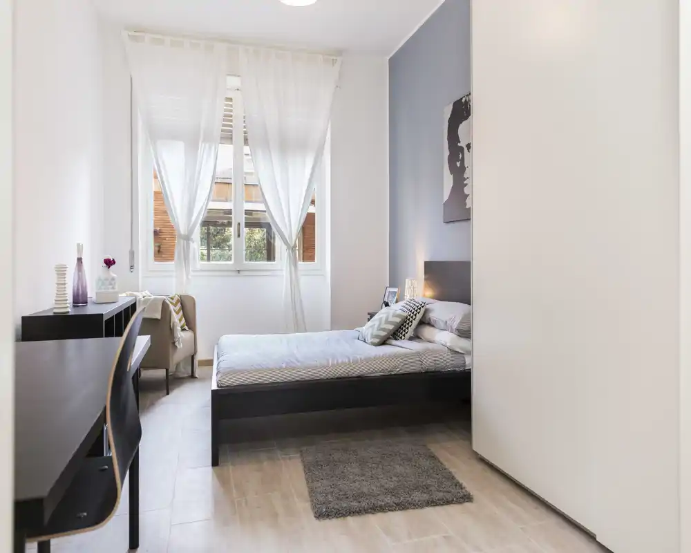 Camera da letto moderna e luminosa con un letto matrimoniale, tessili morbidi e una scrivania vicino alla finestra. La luce naturale rende l'ambiente sereno e arioso.