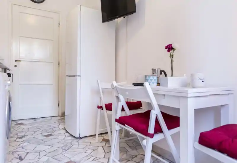 Kitchenette compacte avec une petite table blanche et chaises pliantes aux coussins rouges. Espace lumineux et rangé avec réfrigérateur, TV murale et touches décoratives, adapté à un studio.