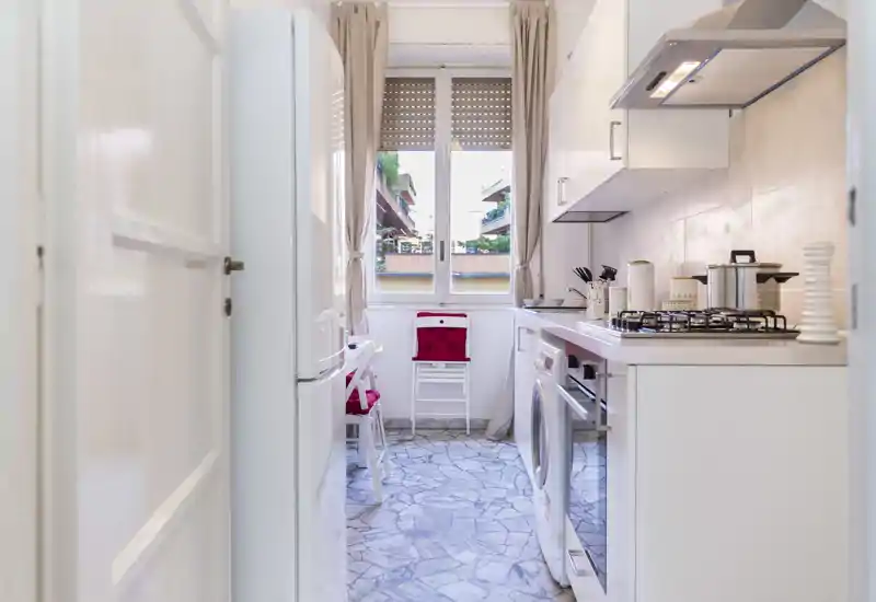 Cuisine en long compacte et lumineuse avec meubles blancs, plaque à gaz, électroménagers et fenêtre apportant de la lumière naturelle.