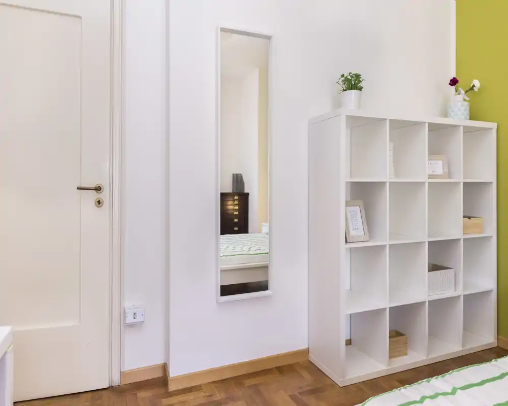 Rincón de dormitorio luminoso y moderno con un espejo estrecho que refleja parte de la cama, una estantería modular blanca y suelo de parquet — estilo minimalista y ordenado, perfecto para anuncios.