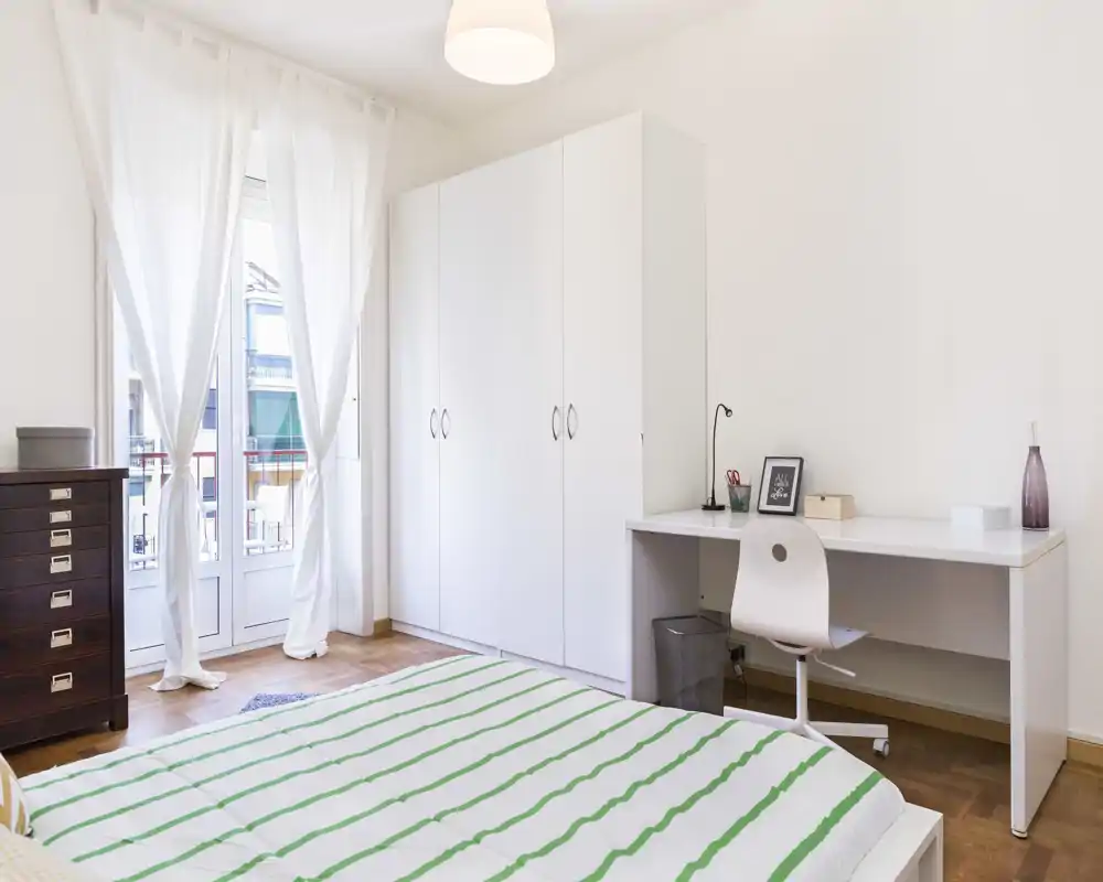 Dormitorio luminoso y minimalista con una cama amplia en primer plano, armario blanco, escritorio y puertas al balcón que inundan la habitación de luz natural.