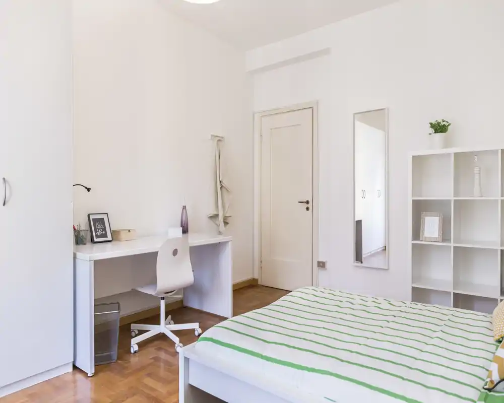 Dormitorio luminoso y minimalista con cama hecha, escritorio y estantería. La luz natural y el suelo de madera clara crean un ambiente tranquilo y aireado, ideal para descansar y trabajar.