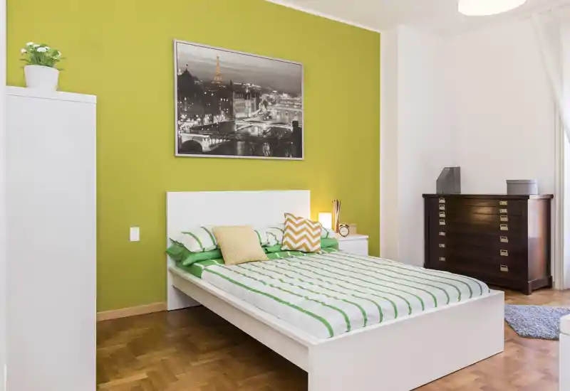 Dormitorio luminoso y bien decorado con cama blanca frente a una pared verde, cojines decorativos y lámpara de noche que crean un ambiente acogedor.