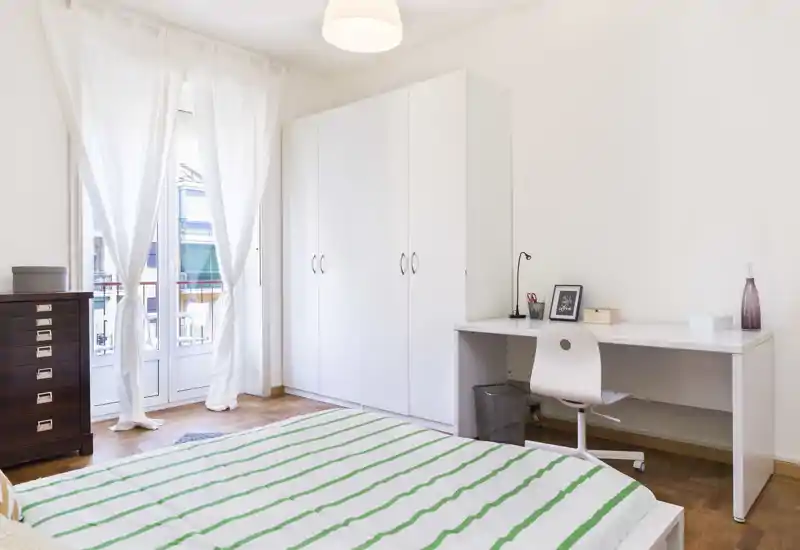 Dormitorio luminoso y minimalista con una cama amplia en primer plano, armario blanco, escritorio y puertas al balcón que inundan la habitación de luz natural.
