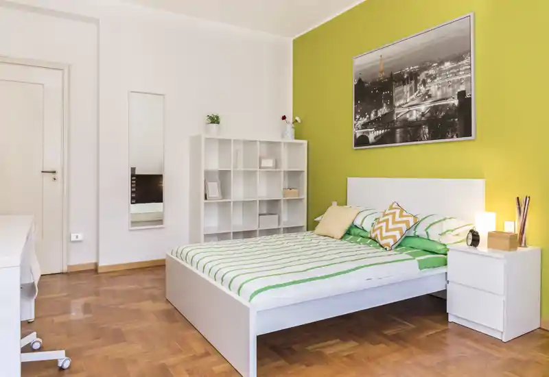 Dormitorio moderno y luminoso con cama blanca, ropa de cama a rayas verdes y cojines decorativos; estantería y mesita de noche minimalistas que aportan calidez.