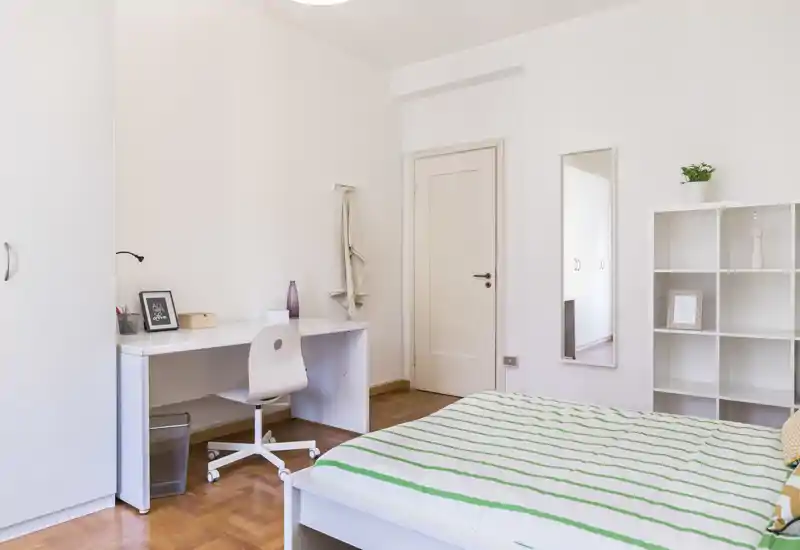 Dormitorio luminoso y minimalista con cama hecha, escritorio y estantería. La luz natural y el suelo de madera clara crean un ambiente tranquilo y aireado, ideal para descansar y trabajar.