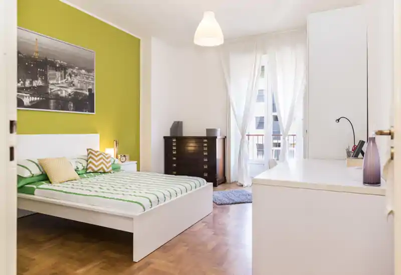 Dormitorio luminoso y bien presentado con cama blanca, pared verde como acento y luz natural desde la ventana/balcón. Cojines decorativos, suelo de parquet y cómoda ordenada ofrecen un ambiente fresco y acogedor, ideal para anuncios.