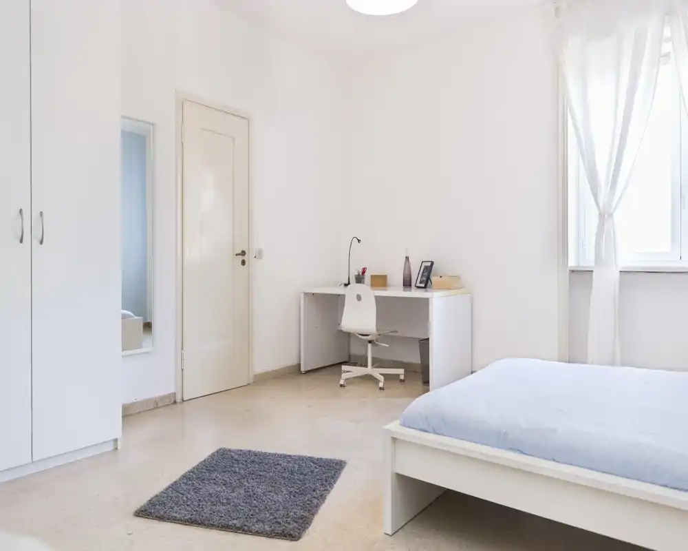 Dormitorio luminoso y minimalista con cama de estructura blanca, ropa de cama azul clara, escritorio y silla junto a la ventana y armario. Ambiente aireado, perfecto para un anuncio que destaque la sencillez y la luz natural.