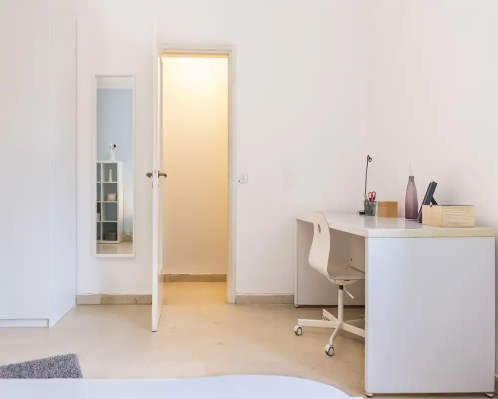 Rincón de dormitorio minimal y luminoso con un escritorio blanco y una pequeña parte de la cama visible en primer plano. El espacio está ordenado y bien iluminado, con un estilo moderno y sencillo.