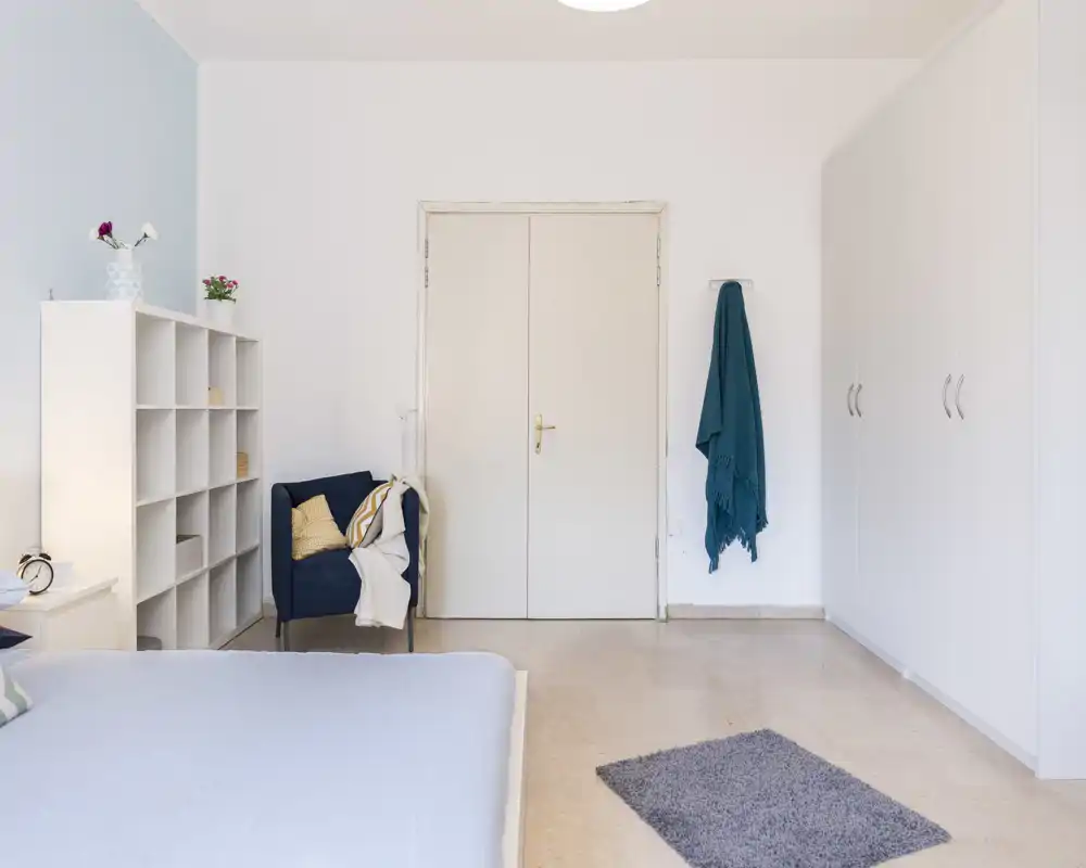 Dormitorio luminoso y minimalista con cama visible, sillón azul, estantería y armarios empotrados; líneas limpias y textiles suaves para un ambiente tranquilo.