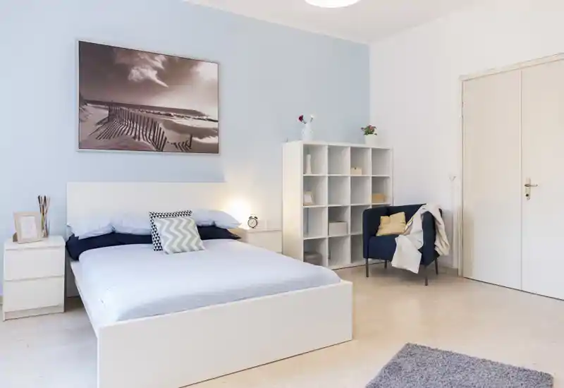 Dormitorio luminoso y bien decorado con cama blanca, mesitas, estantería modular y un cómodo sillón — perfecto para fotos de alquiler que muestran confort y almacenamiento.