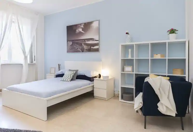 Dormitorio luminoso y aireado con cama doble blanca, mesitas y un sillón acogedor; paredes azul claro y luz natural que crean un ambiente sereno y acogedor.