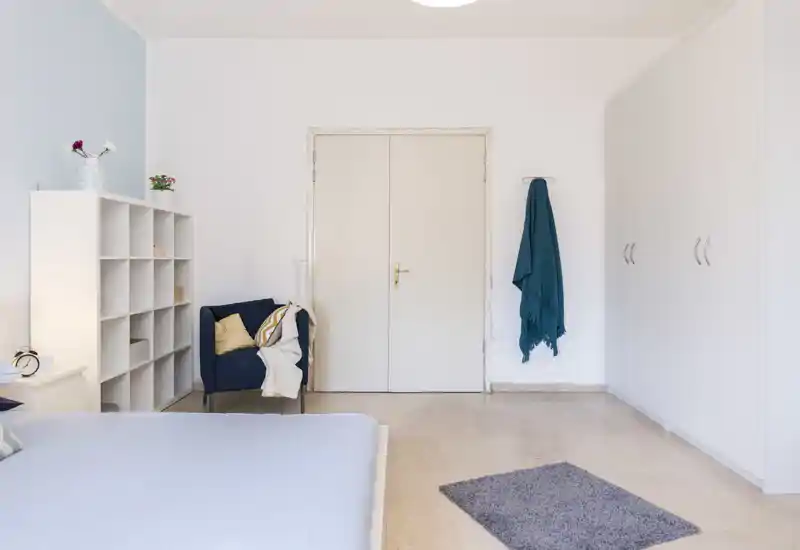 Dormitorio luminoso y minimalista con cama visible, sillón azul, estantería y armarios empotrados; líneas limpias y textiles suaves para un ambiente tranquilo.