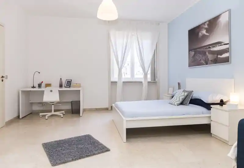 Dormitorio luminoso y minimalista con cama doble, mesita con lámpara y un pequeño escritorio junto a la ventana. Tonos neutros y pared azul crean un ambiente tranquilo y luminoso, perfecto para anuncios.