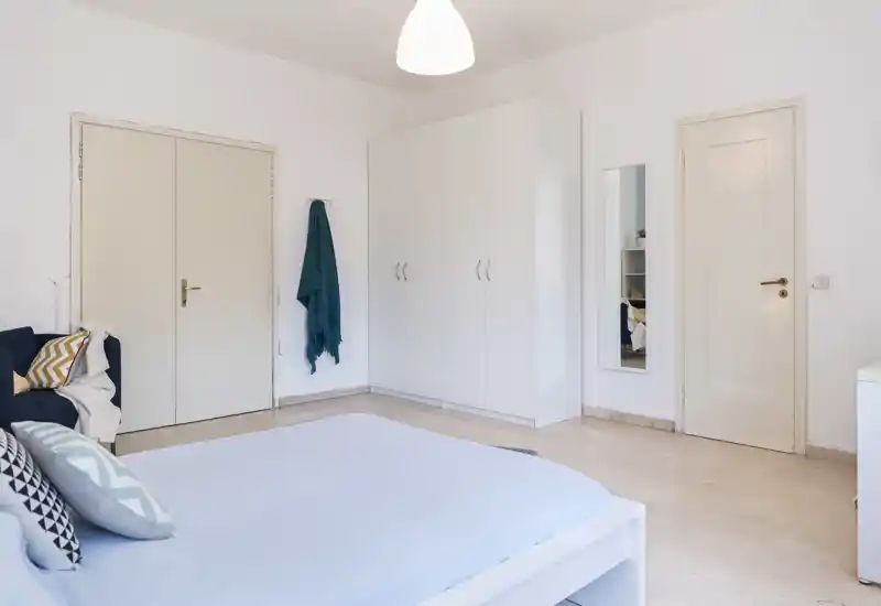 Dormitorio luminoso y minimalista con cama doble en primer plano, armario blanco, espejo de cuerpo entero y cojines que aportan comodidad.
