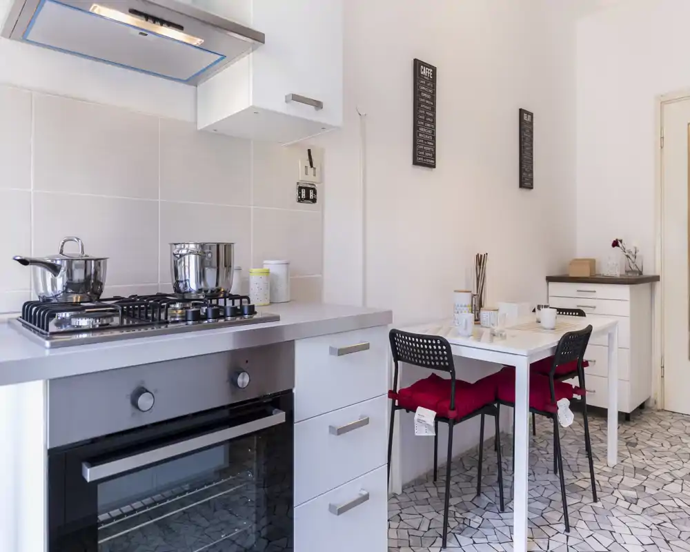 Cocina moderna y compacta con placa de gas y horno integrado, pequeña mesa con dos sillas y cojines rojos — luminosa, funcional y ordenada para una comida acogedora.