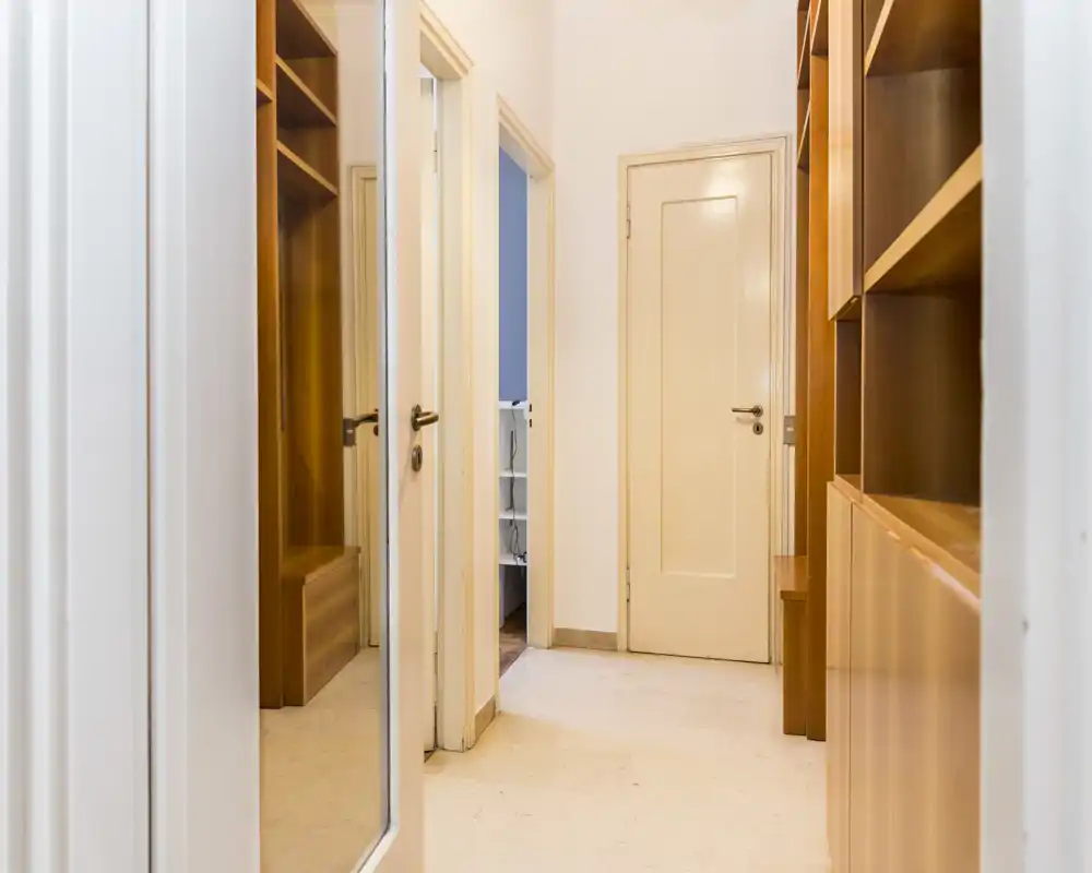 Recibidor/pasillo luminoso con estanterías de madera empotradas y armario con espejo, suelo de baldosas limpio y puerta interior cerrada.