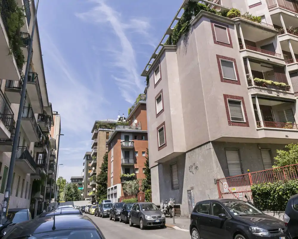 Vista a nivel de calle de una manzana residencial con coches aparcados y edificios de pisos bajo un cielo azul.