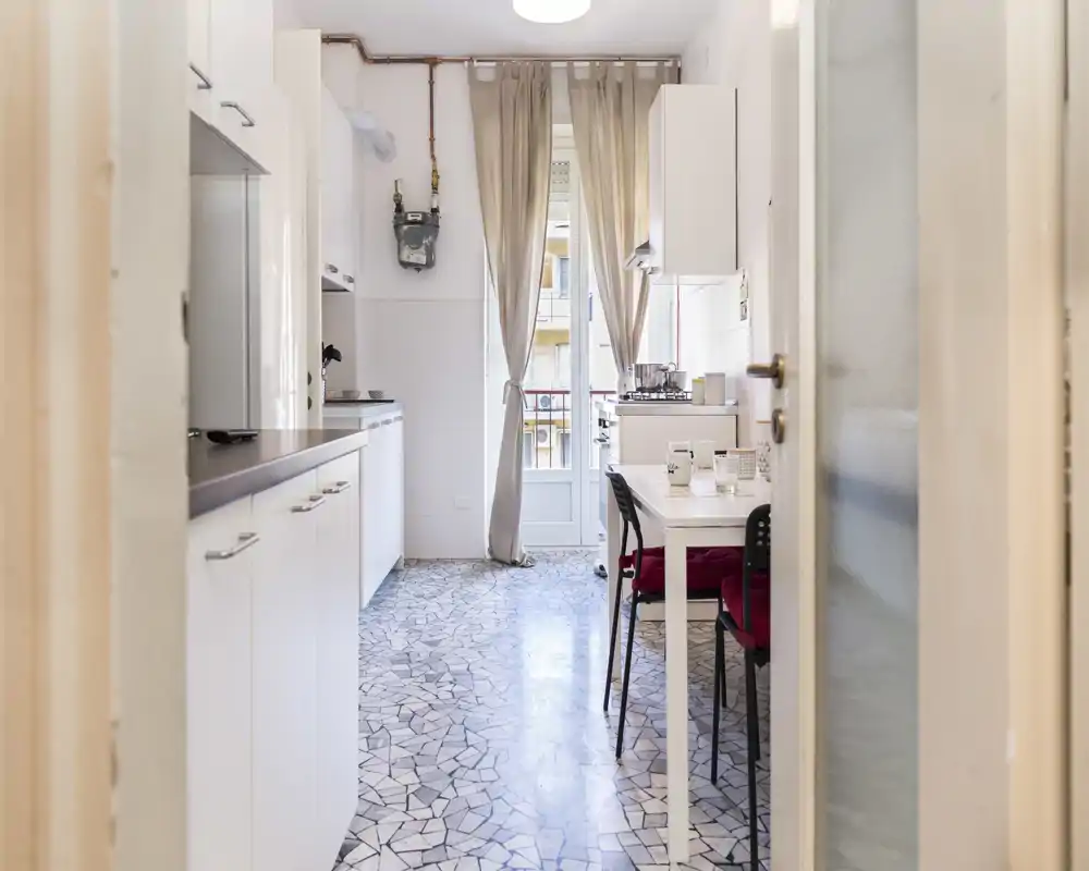 Cocina luminosa y estrecha con armarios blancos, pequeña mesa de comedor y luz natural desde la puerta del balcón — práctica y bien cuidada.