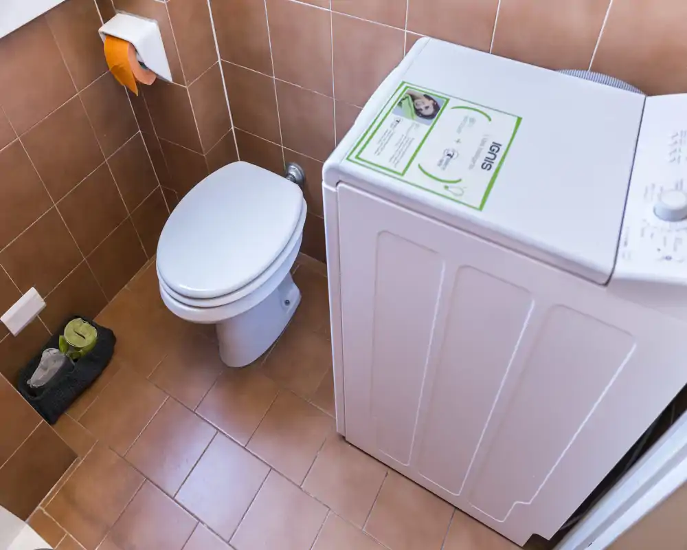 Baño pequeño con inodoro y lavadora de carga superior sobre baldosas color terracota; distribución compacta y limpia, adecuada para un piso.