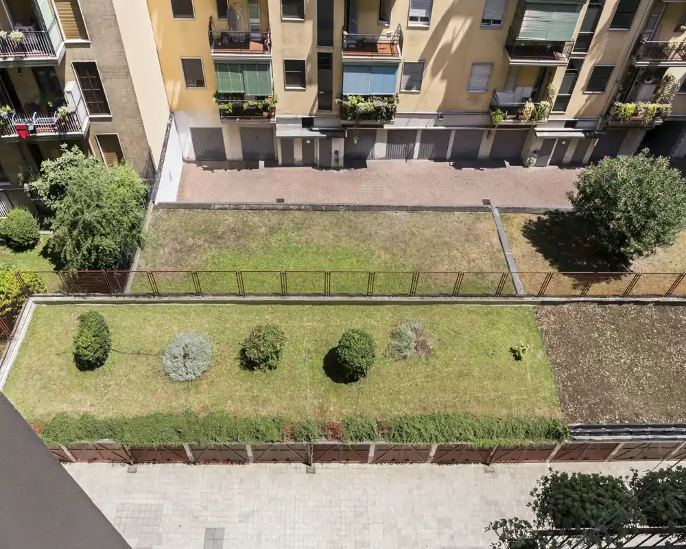 Patio comunitario soleado con franja de césped, pequeños arbustos y vallas — ideal para mostrar el espacio exterior compartido y la vegetación.