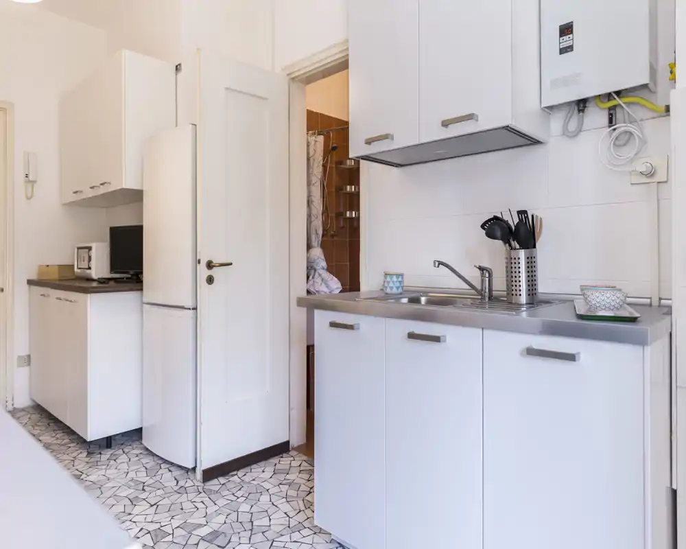 Cocina compacta y moderna con armarios blancos, fregadero de acero inoxidable y utensilios ordenados sobre la encimera. Espacio luminoso y bien organizado, ideal para estancias cortas.