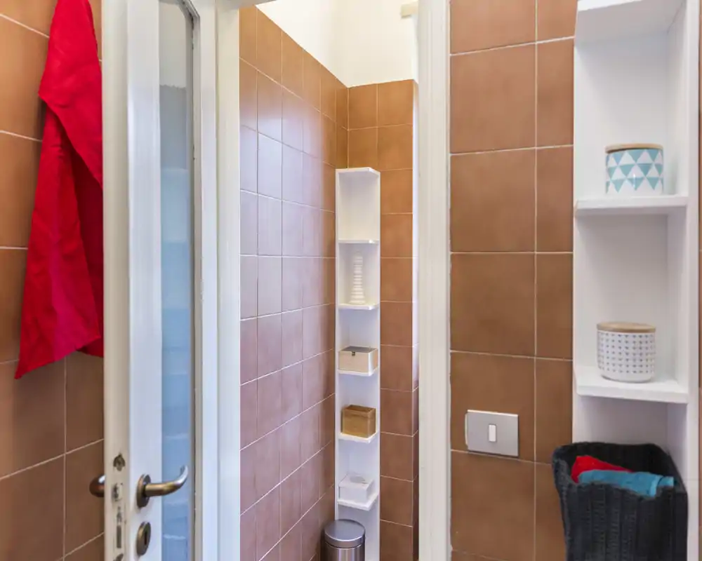 Baño compacto con azulejos color terracota, estantería alta y estrecha para artículos y pequeño cubo de basura; ordenado y bien iluminado.