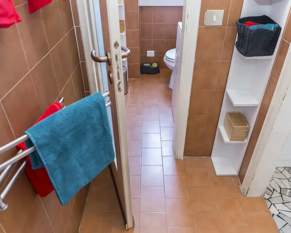 Baño pequeño con azulejos, inodoro visible, estantería y toallas coloridas; espacio luminoso y ordenado, adecuado para alquileres de corta estancia.