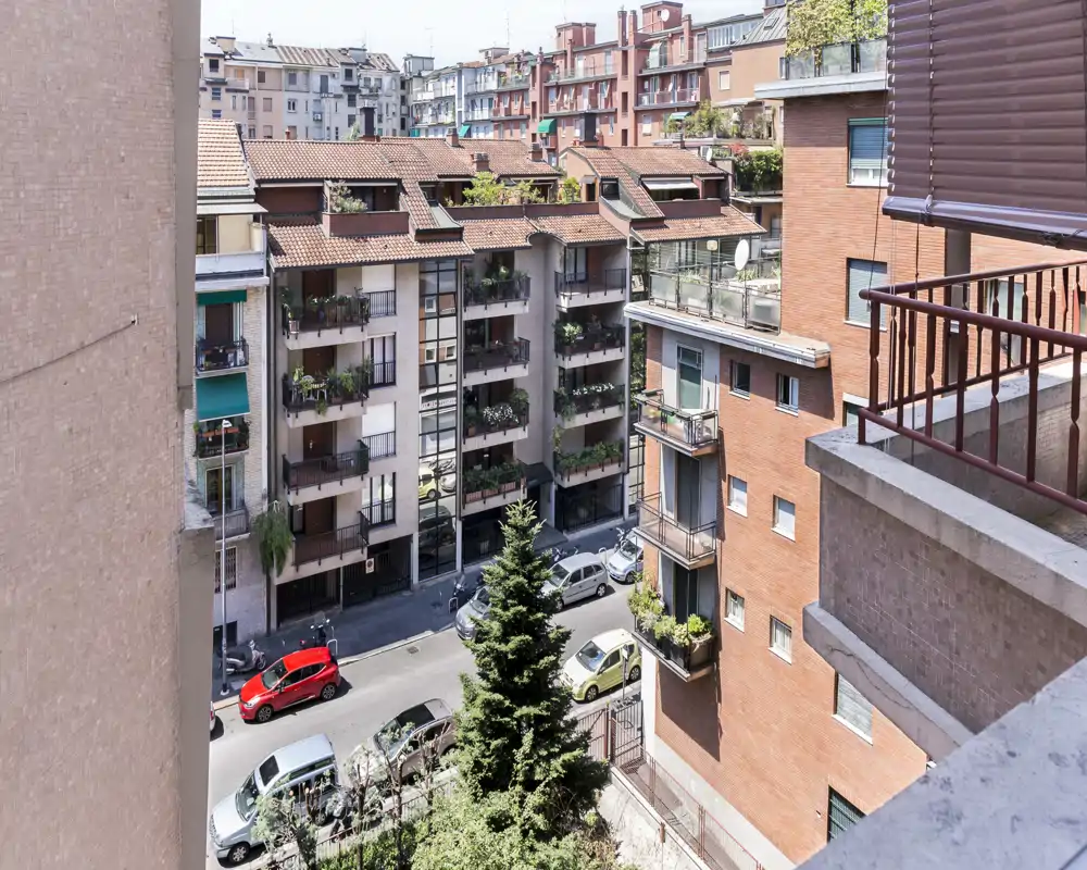 Vista desde un balcón urbano hacia una calle residencial y edificios contiguos, con macetas y coches aparcados visibles — ideal para mostrar el entorno urbano y la luz natural.