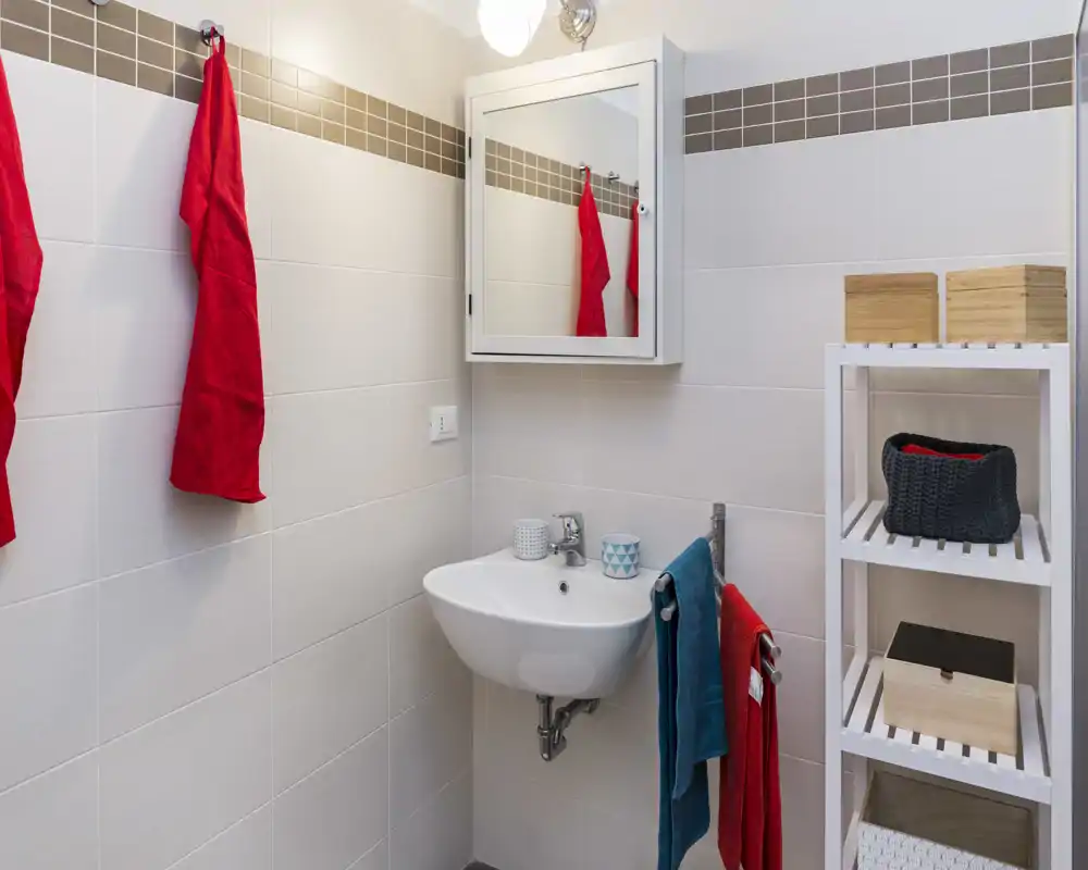Baño compacto y moderno con lavabo suspendido, armario con espejo y estantería abierta. Azulejos neutros y toallas coloridas crean un aspecto limpio y luminoso.