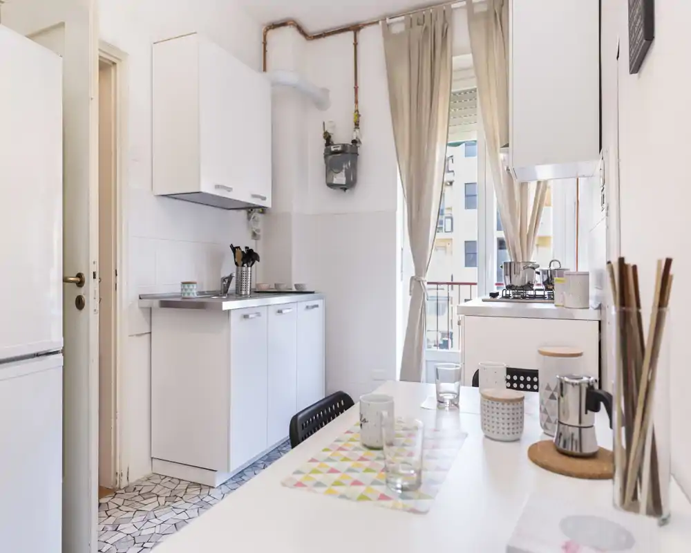 Cocina compacta y luminosa con muebles blancos, pequeña placa junto a la ventana y mesa ordenada con tazas y manteles — ideal para un apartamento urbano acogedor.