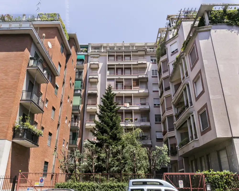 Vista exterior de un bloque de apartamentos residenciales con balcones, un pequeño patio ajardinado y coches aparcados; imagen diurna clara adecuada para mostrar la fachada y la zona exterior común.