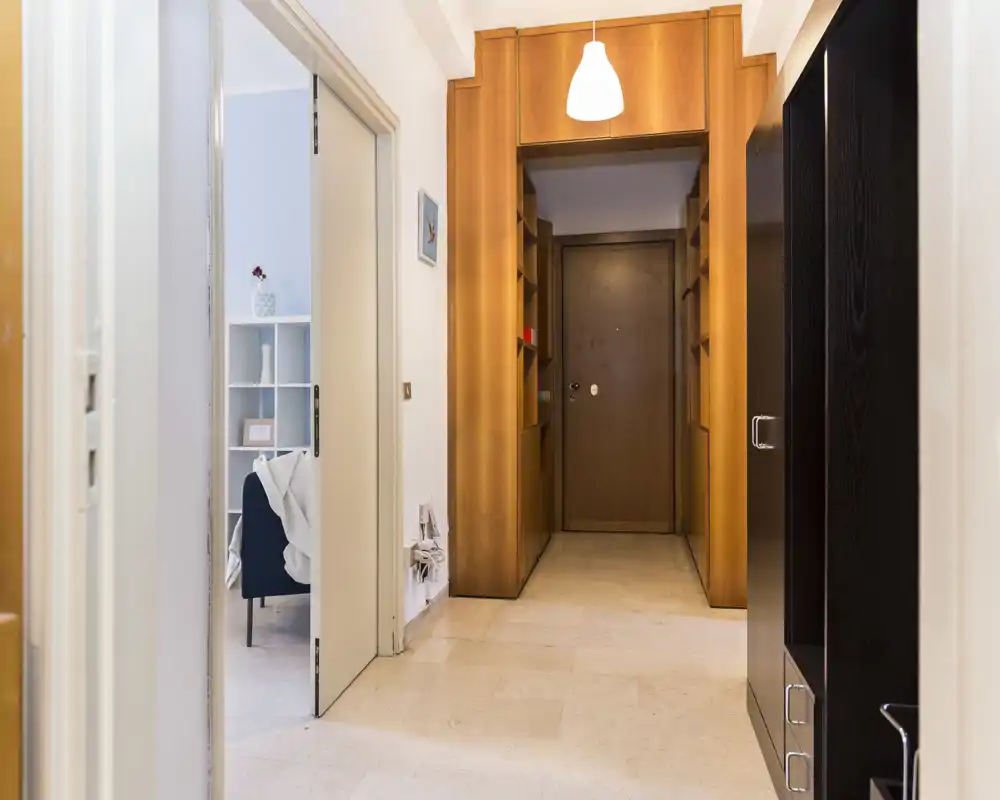 Vestíbulo de entrada luminoso y bien cuidado con estanterías empotradas de madera y puerta principal segura; suelo de piedra y lámpara cálida crean una sensación acogedora.