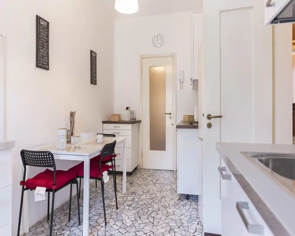Cocina compacta y luminosa con una pequeña mesa de comedor y dos sillas, muebles blancos modernos y suelo de mosaico — ideal para un piso en la ciudad.