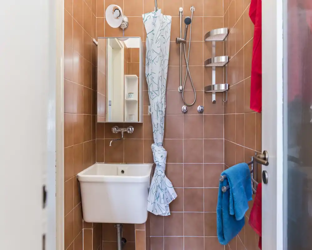 Baño compacto con azulejos, lavabo mural, ducha de mano, cortina y estantes en la pared; espacio luminoso y bien iluminado, ideal para un apartamento pequeño.