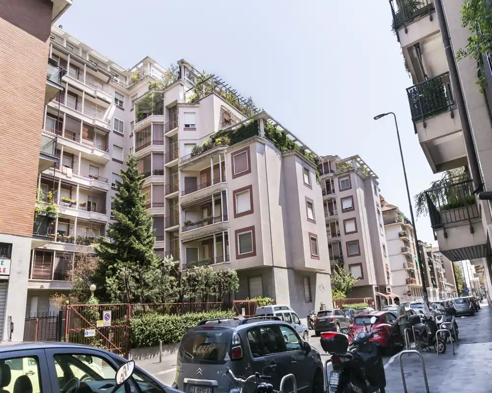 Vista de calle luminosa que muestra la fachada de un edificio residencial de varias plantas con balcones, coches y motos aparcados en la acera.