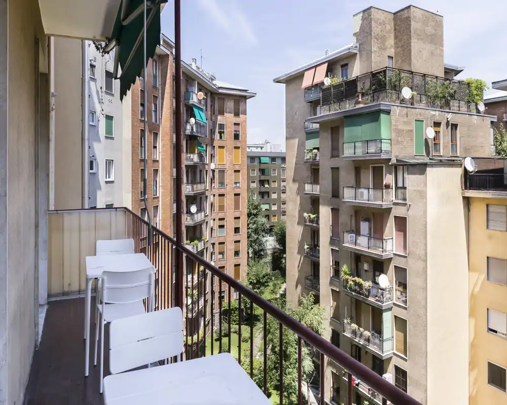 Balcón privado soleado con asientos exteriores y vistas a los edificios residenciales y un pequeño patio.