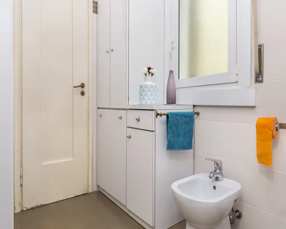 Baño compacto y limpio con bidé, armarios empotrados y ventana translúcida; toallas de colores que aportan contraste y una sensación moderna y ordenada.