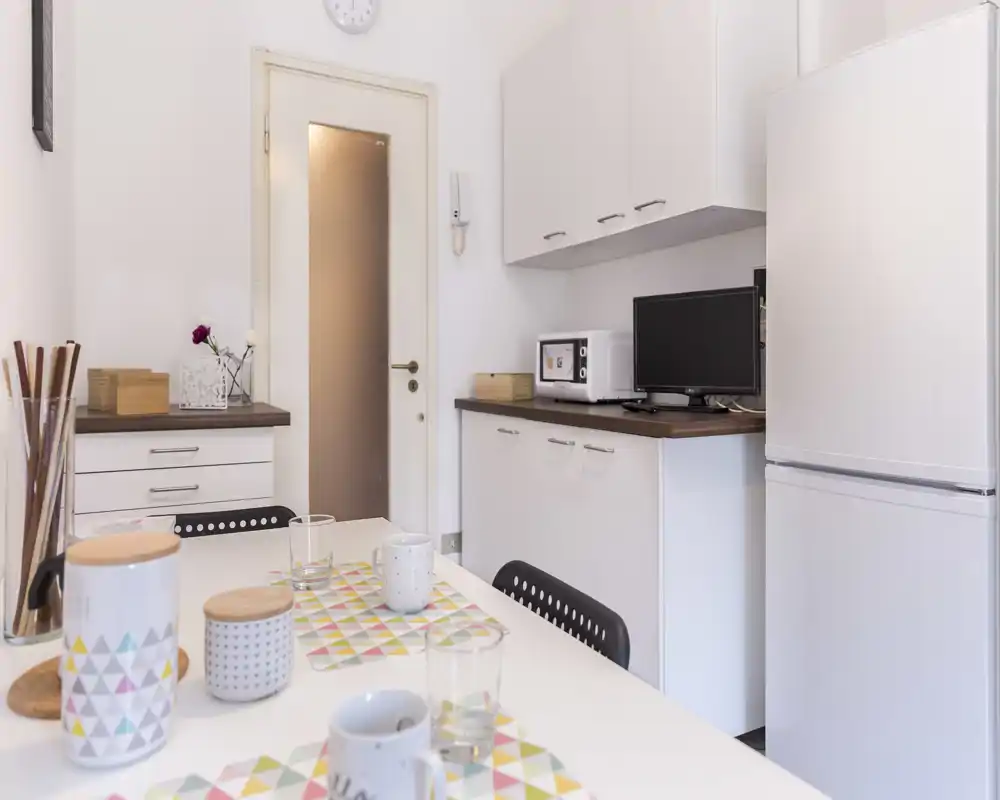 Rincón de cocina luminoso y compacto con mesa en primer plano, frigorífico y pequeños electrodomésticos visibles; muebles modernos y puesta de mesa informal.