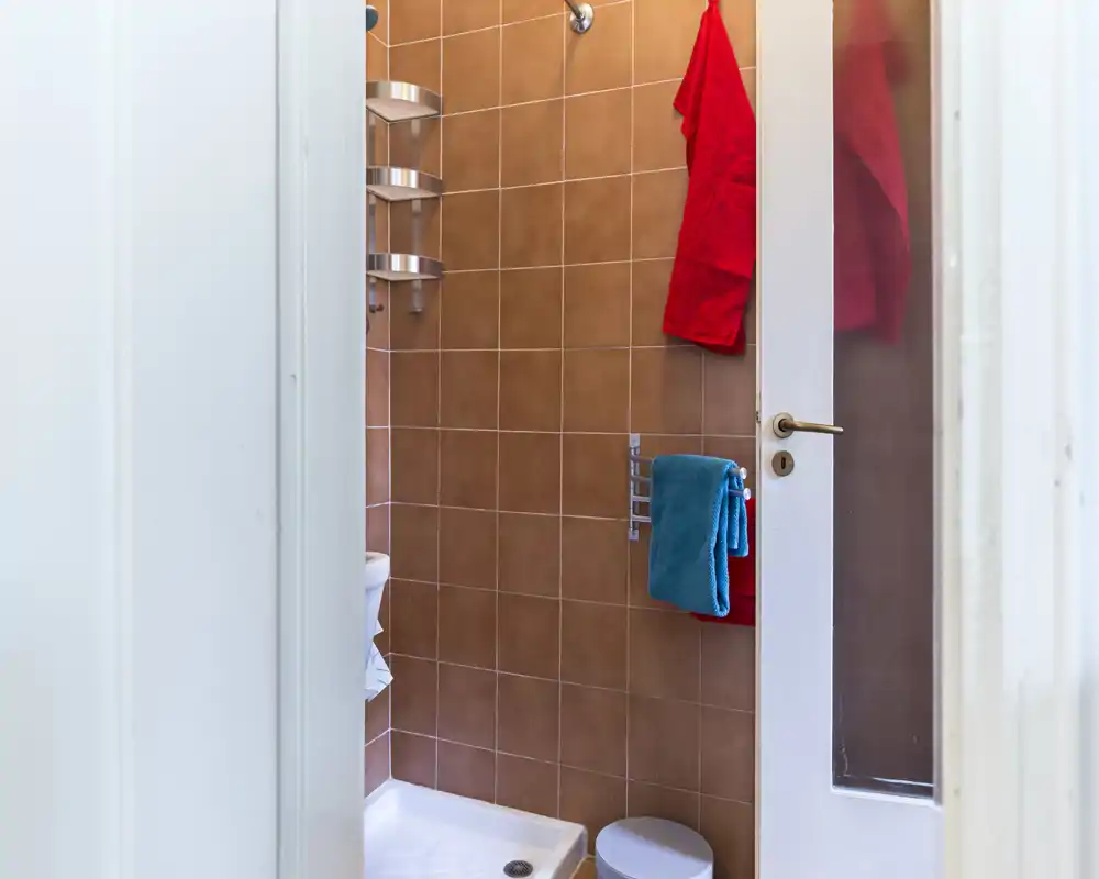 Baño compacto con esquina de ducha con azulejos, perchas de pared y pequeño taburete — luminoso, ordenado y listo para usar.