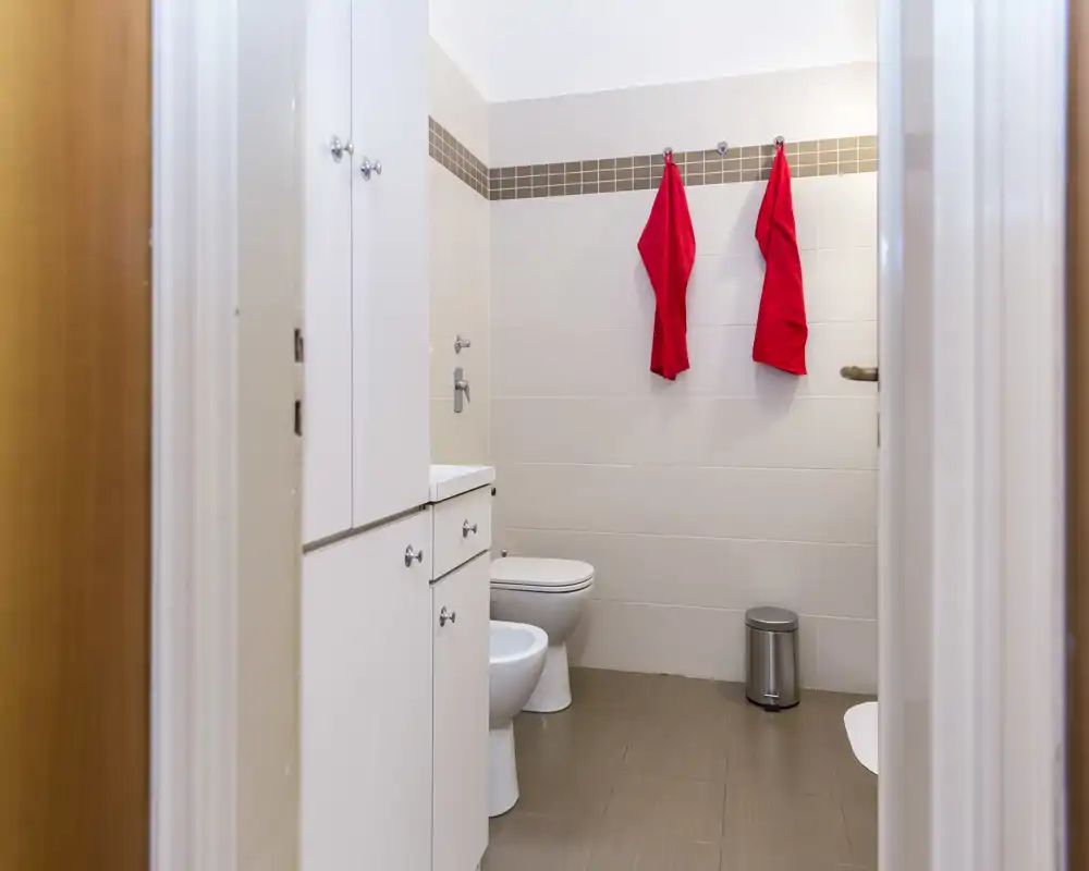 Baño moderno y limpio con dos toallas rojas colgadas, bidé y váter, mueble blanco y azulejos neutros: luminoso y bien cuidado.