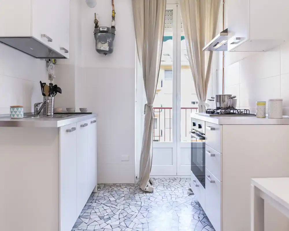 Cocina moderna y luminosa con armarios blancos, cocina de gas y puerta al balcón. Cortinas ligeras y suelo de baldosas con patrón crean un ambiente acogedor.