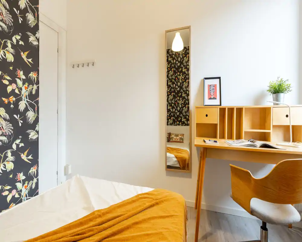 Bright, styled bedroom with a visible bed partly shown, warm ochre throw and a wooden desk by the window; tasteful wallpaper adds character.