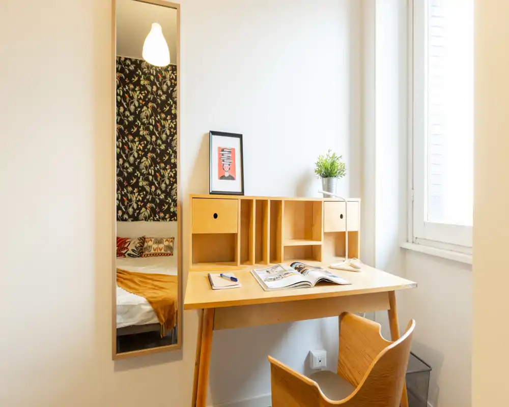 Compact bedroom corner with a wooden desk, bright window light and a partial view of the bed reflected in a tall mirror. Cozy textiles and patterned wallpaper are visible, creating a warm, inviting atmosphere.