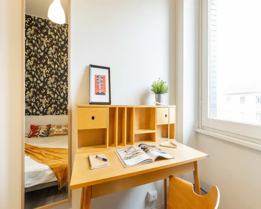 Bright, cozy bedroom corner with a wooden desk and a partially visible bed framed by patterned wallpaper—ideal for a single tenant or student.