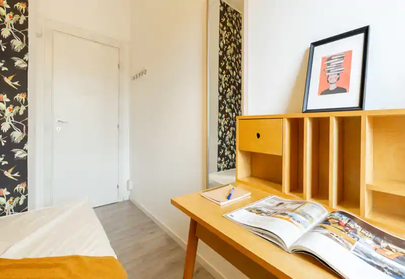 Bright, compact bedroom corner with a wooden desk and a partially visible bed; tasteful floral wallpaper and mirror add character.